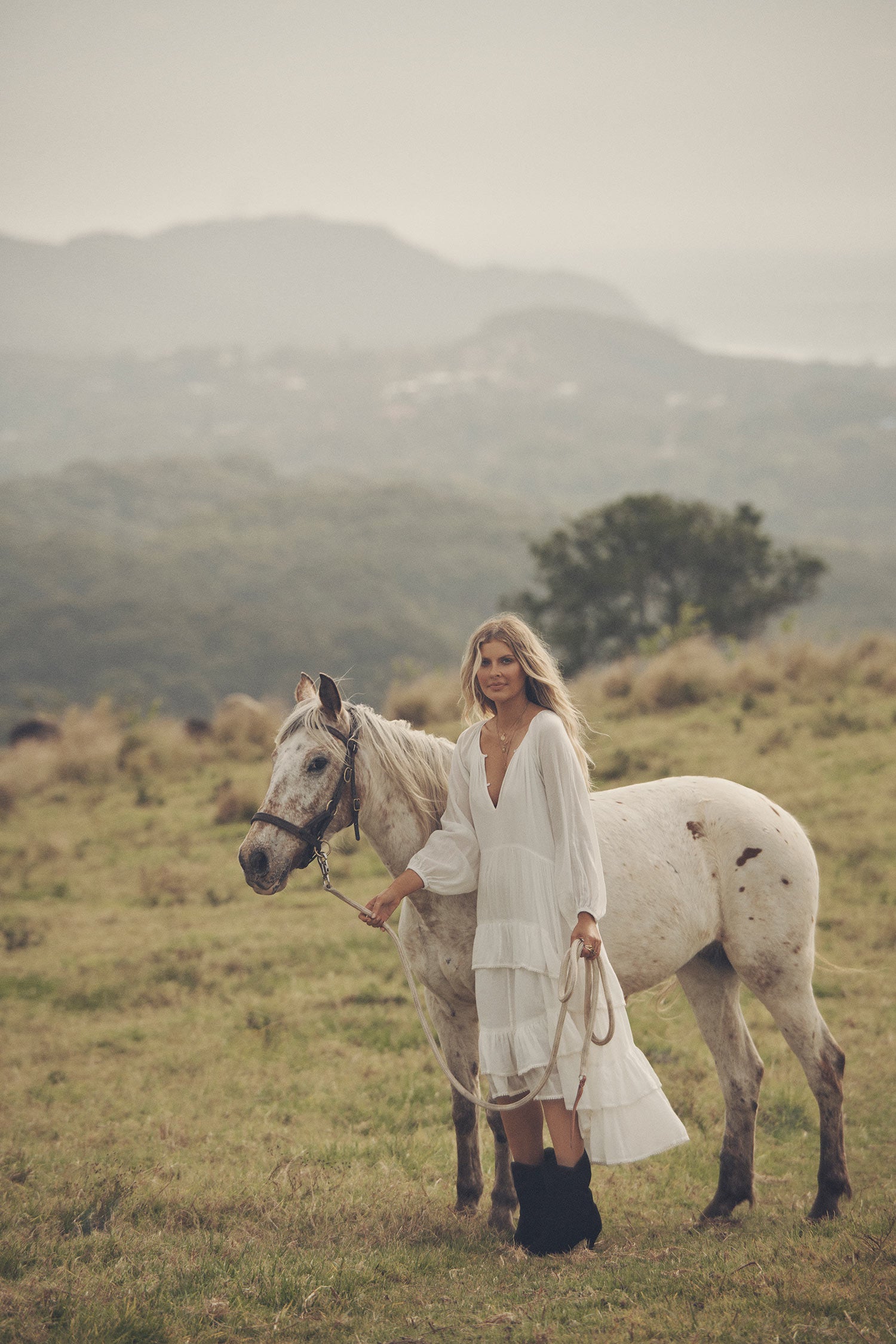 Influencer and model, Elle Ferguson, posing in a green pasture with a white horse while wearing gold jewellery, the Spell Cult Suede Slouch Boots with the white, tiered Moonflower Gown featuring frills and elasticated cuffs. 