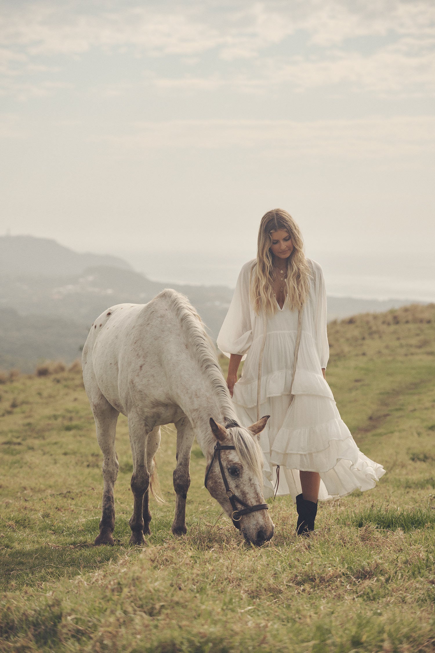 Influencer and model, Elle Ferguson, posing in a green pasture with a white horse while wearing gold jewellery, the Spell Cult Suede Slouch Boots with the white, tiered Moonflower Gown featuring frills and elasticated cuffs. 