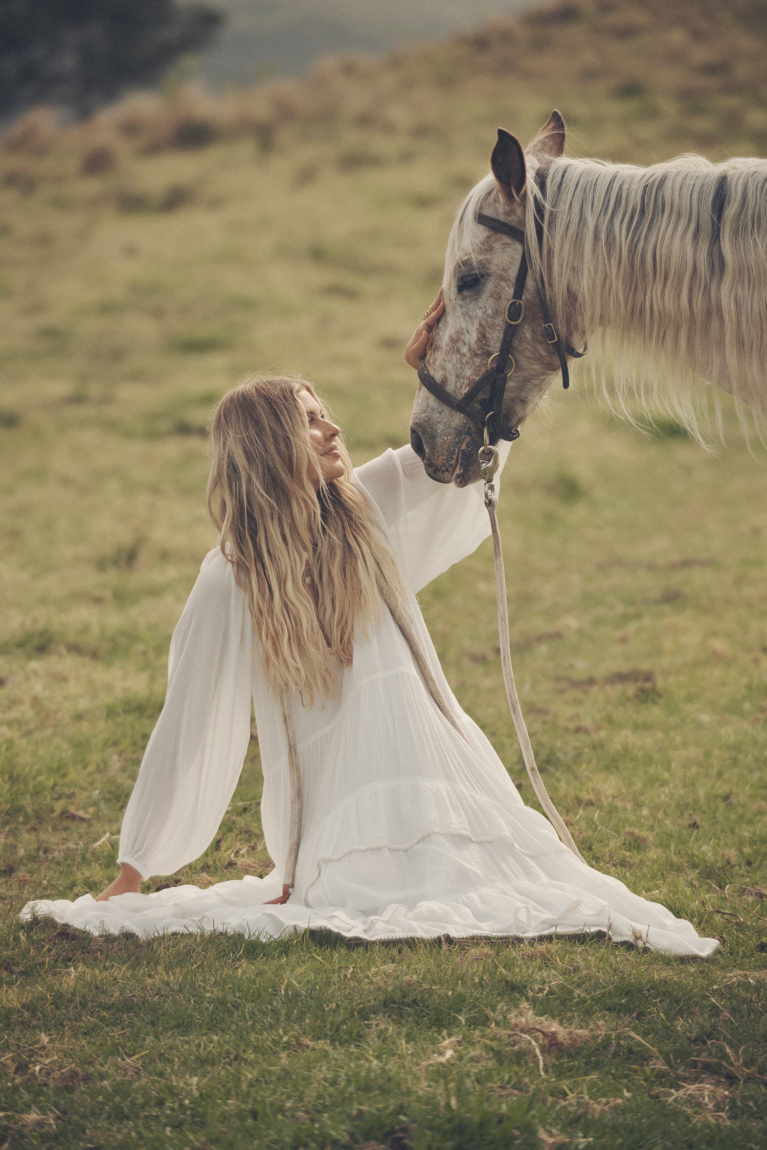 Influencer and model, Elle Ferguson, posing in a green pasture with a white horse while wearing gold jewellery, the Spell Cult Suede Slouch Boots with the white, tiered Moonflower Gown featuring frills and elasticated cuffs. 