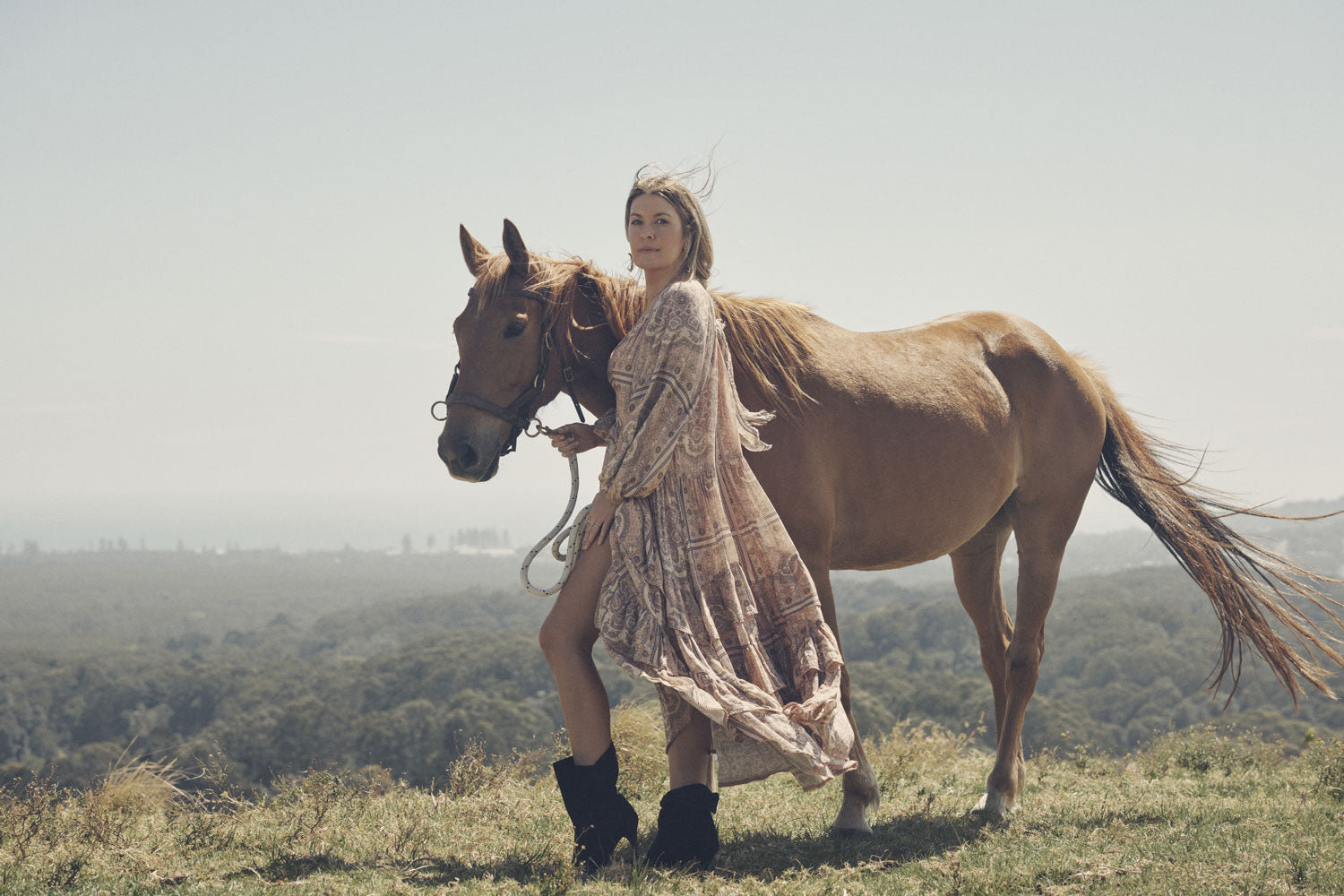 Jeweller and campaign model, Lucie Ferguson, posing in a green pasture with a brown horse while wearing gold jewellery, the Spell Cult Suede Slouch Boots with the pale pink, tiered Rumour Print Elle Gown featuring frills and elasticated cuffs.
