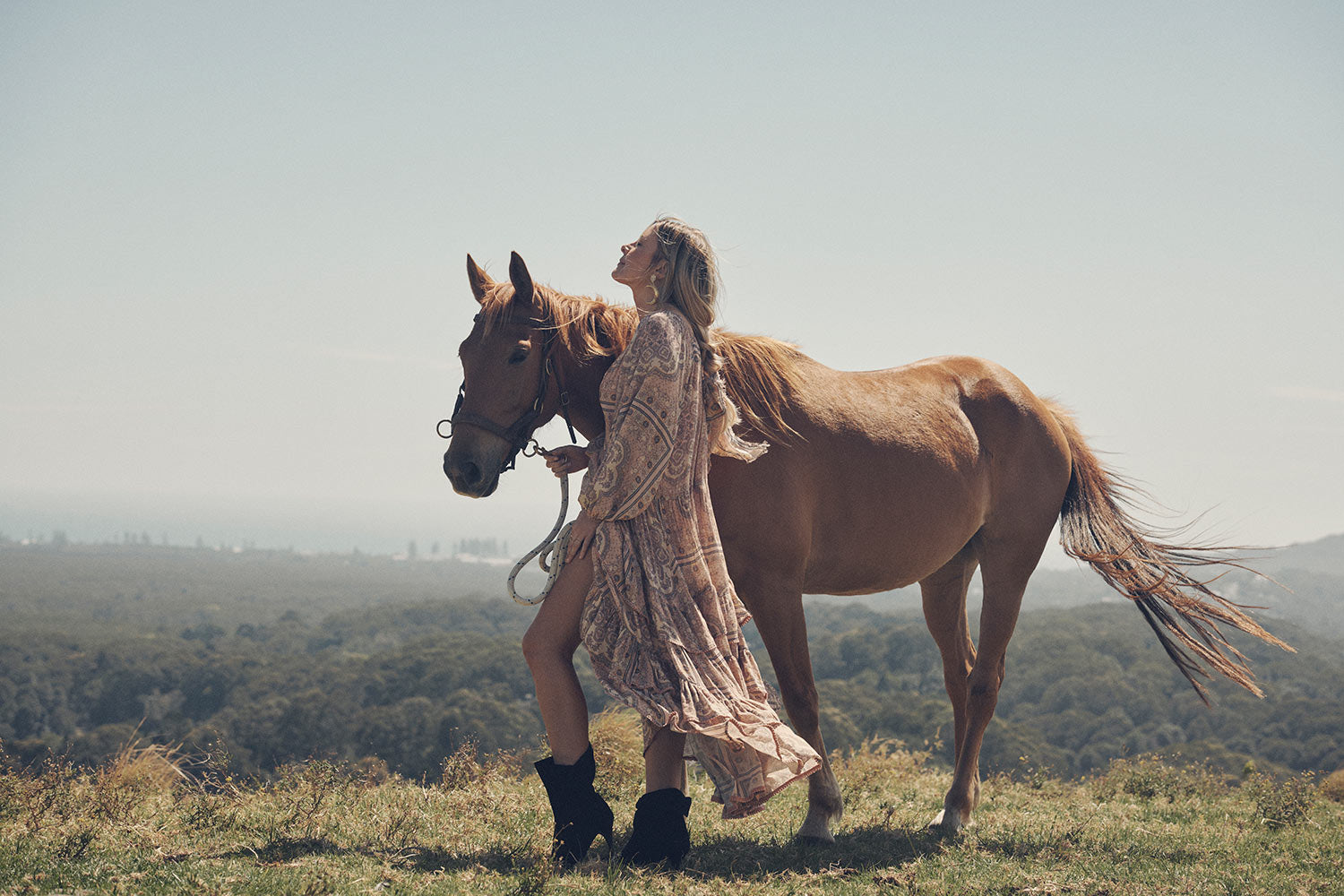 Jeweller and campaign model, Lucie Ferguson, posing in a green pasture with a brown horse while wearing gold jewellery, the Spell Cult Suede Slouch Boots with the pale pink, tiered Rumour Print Elle Gown featuring frills and elasticated cuffs.