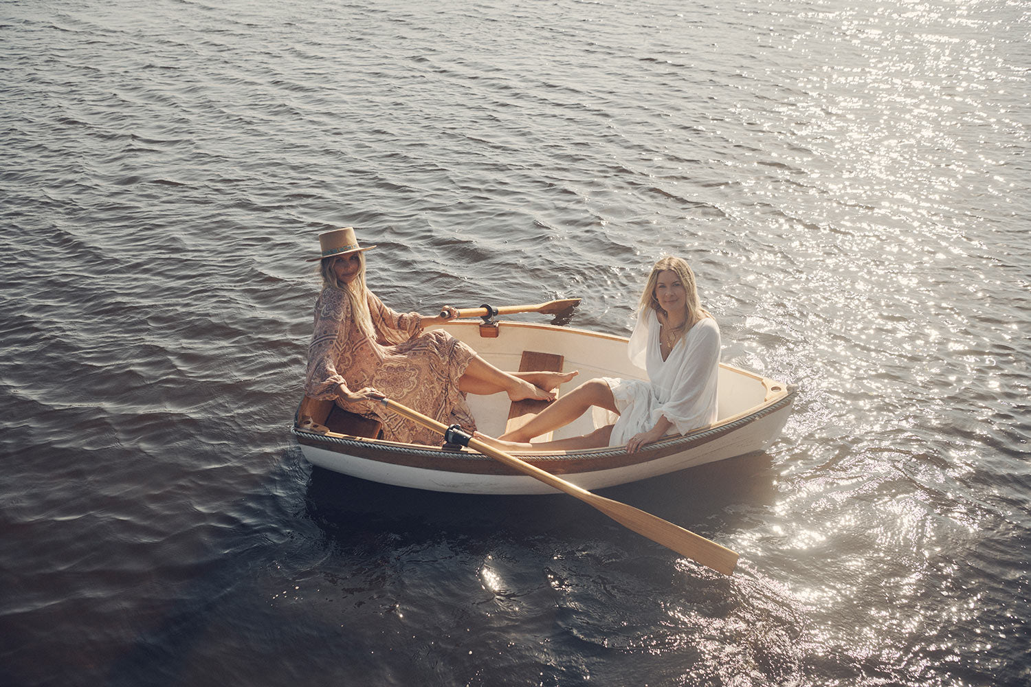 Sisters, Lucie and Elle Ferguson, on a white rowboat in the middle of a river wearing the white tiered Spell Moonflower Gown and the pale pink Spell Rumour Print Elle Gown, both featuring tiered skirts, ruffles and elasticated cuffs. 