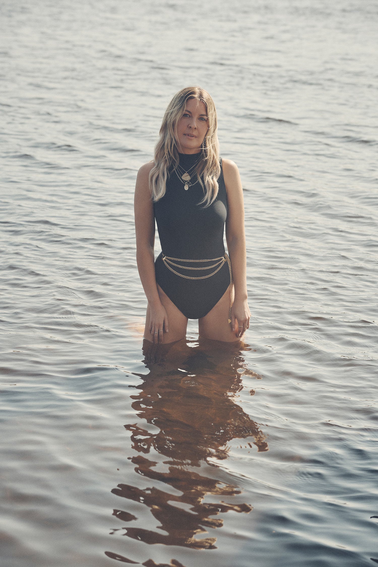 Jeweller and campaign model, Lucie Ferguson, standing in the shallows wearing a black one-piece swimsuit paired with gold necklaces and the brass Spell Belladonna Chain Belt, featuring gold chain links and moon detailing. 