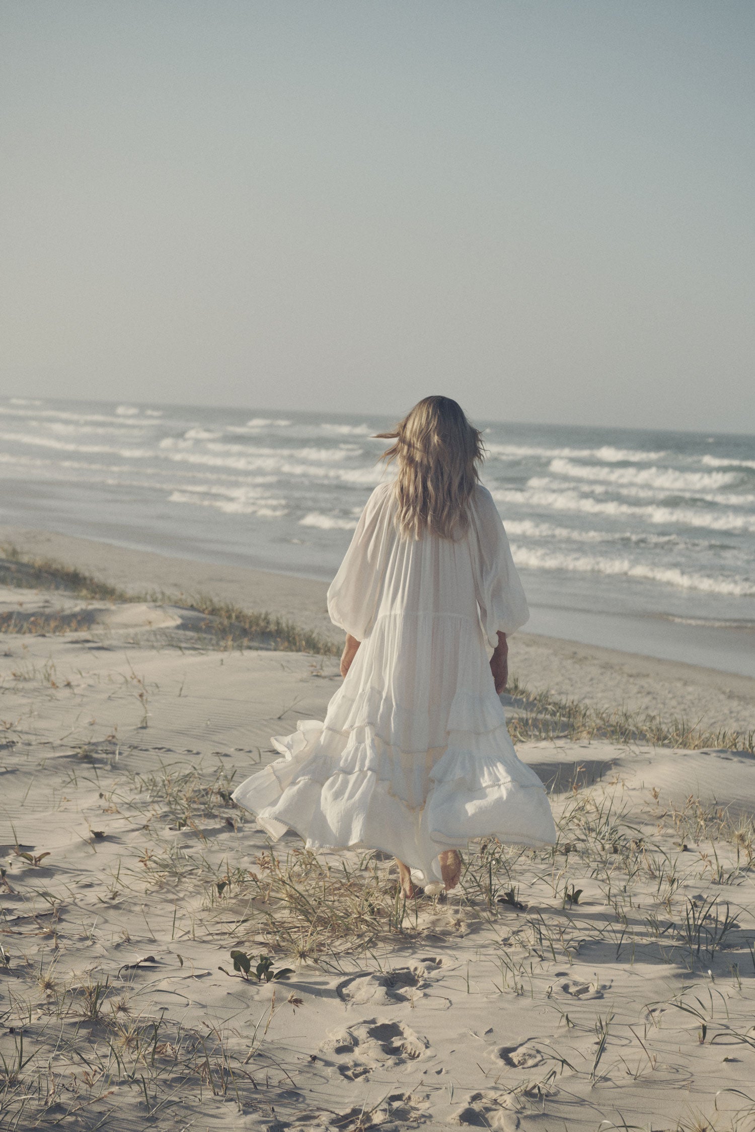  Jeweller and campaign model, Lucie Ferguson, walking on a sand dune while wearing gold jewellery and the white Spell Moonflower Gown featuring elasticated cuffs and a tiered skirt. 