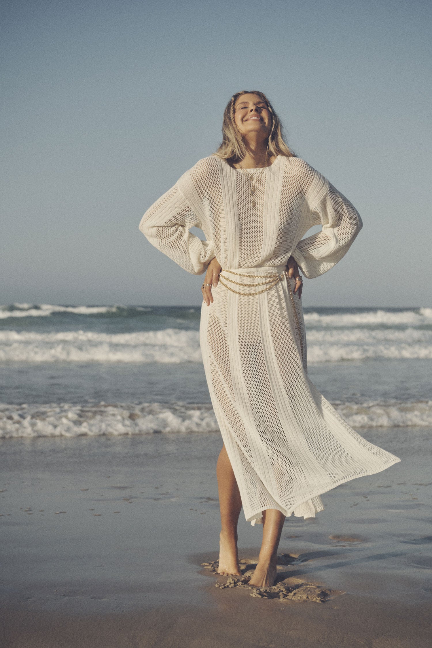 Influencer and model, Elle Ferguson, posing on a beach while wearing gold jewellery, the Spell Belladonna Chain Belt and the cream Rush Knit Midi Dress, featuring side splits and an elasticated waistline. 