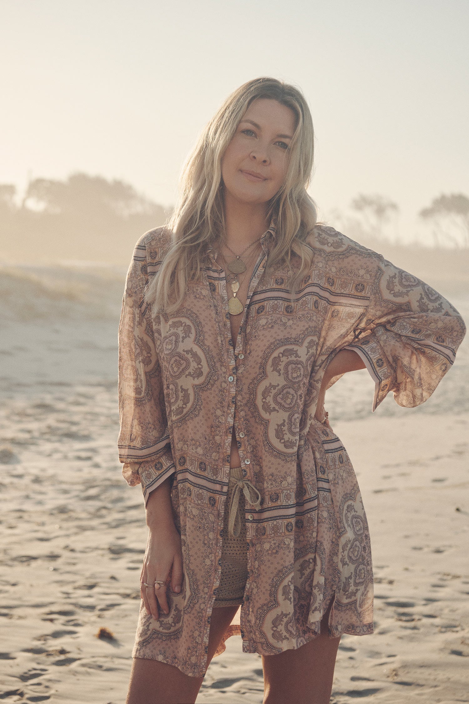 Jeweller and campaign model, Lucie Ferguson, posing on a beach while wearing gold jewellery, the golden Spell Moonlight shorts and the pale pink Spell Rumour Print Shirt Dress, featuring a shell button down front and collar. 