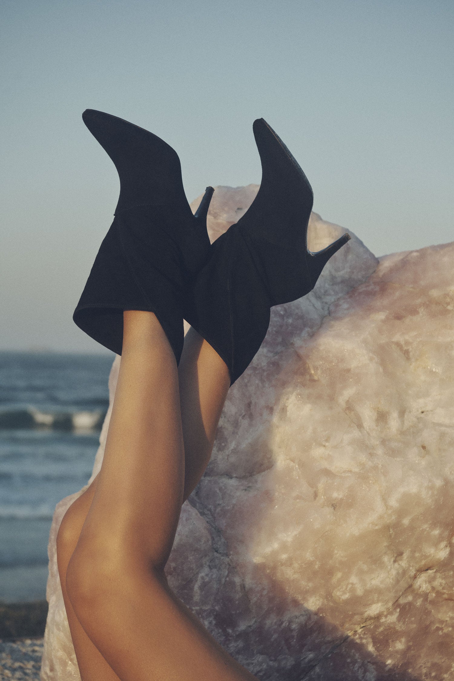 A close- up image of the Spell black Cult Suede Boots, featuring a pointed toe and narrow heel, with a crystal and beach in the background. 