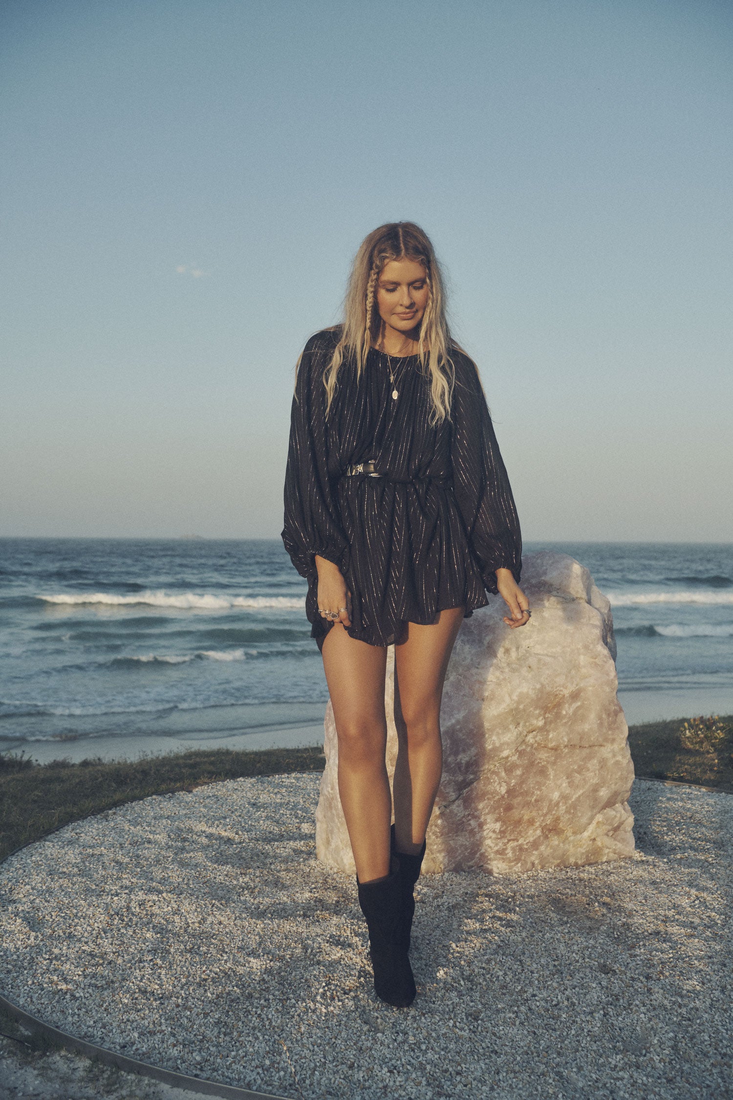 Influencer and model, Elle Ferguson, posing in front of a large crystal with the beach in the background while wearing gold jewellery, a black belt, the Spell Cult Suede Slouch Boots and the Spell black, metallic Jagger Mini Dress with elasticated cuffs. 