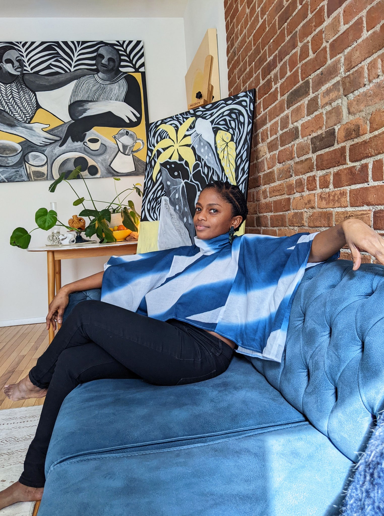 Portrait of Nathalie Jolivert reclining on blue sofa with her paintings hung on brick wall