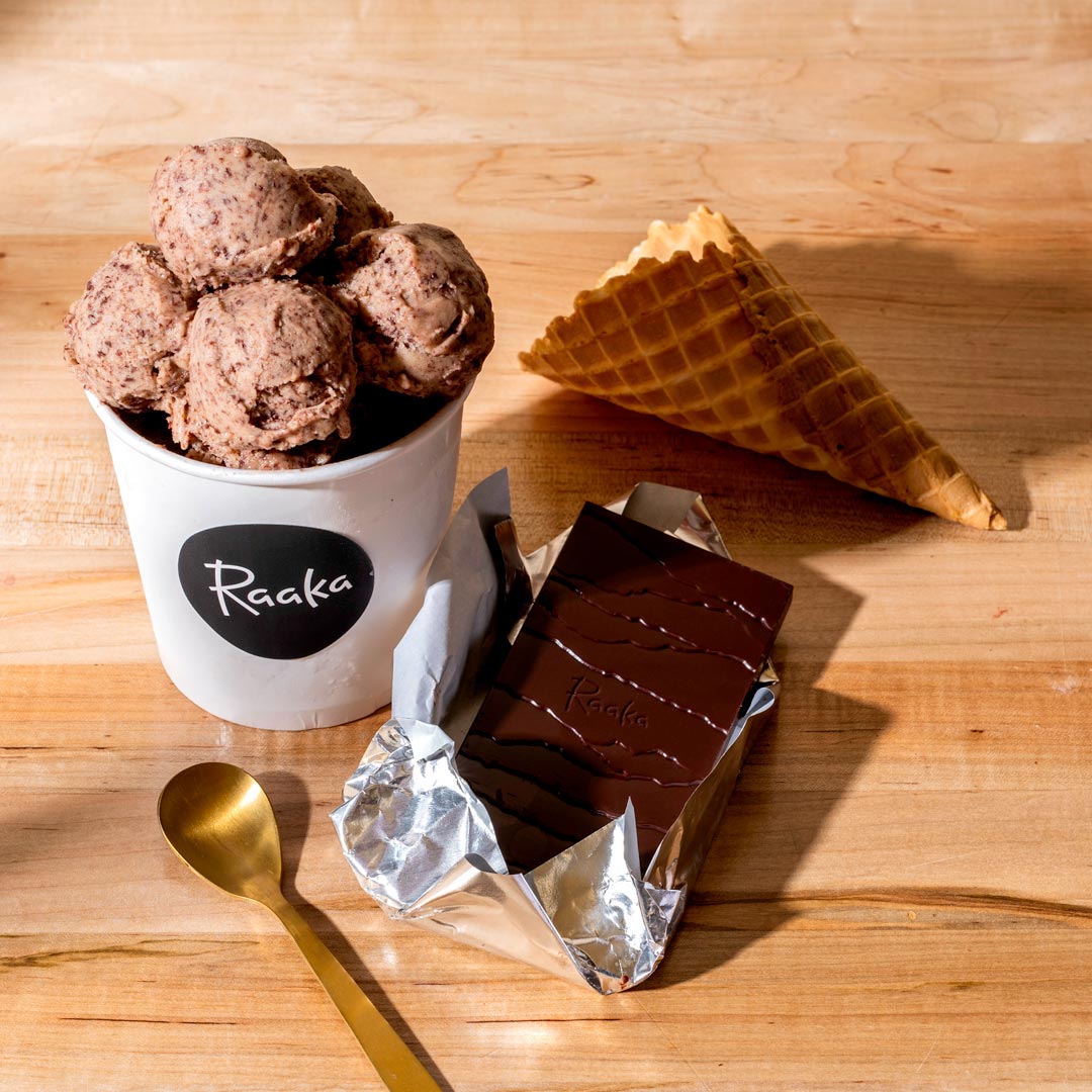 A cup of chocolate chip ice cream, Raaka chocolate bar, waffle cone, and golden spoon on a wooden table.