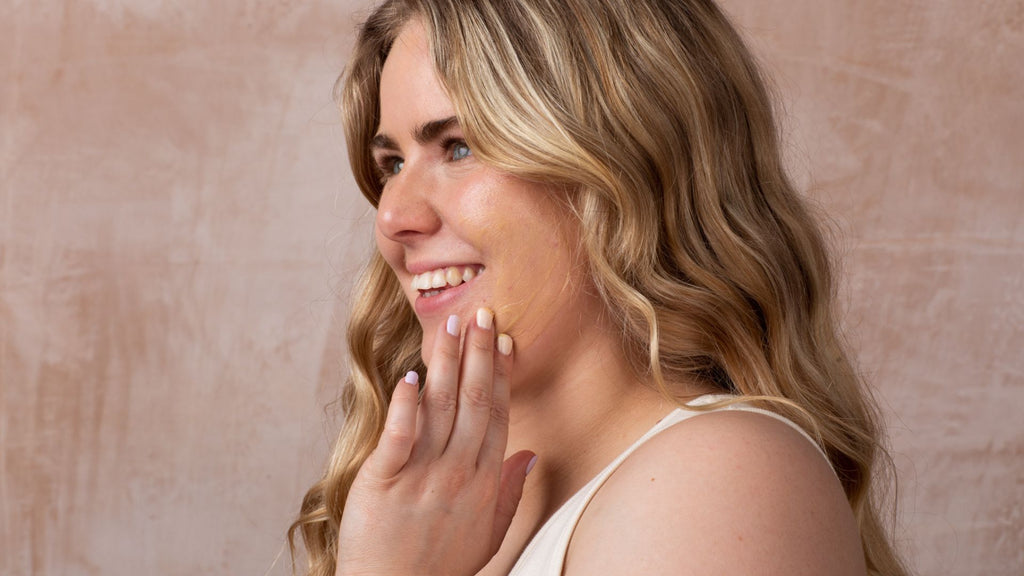 young female with wavy blonde hair smiling whilst applying moisturiser to her face