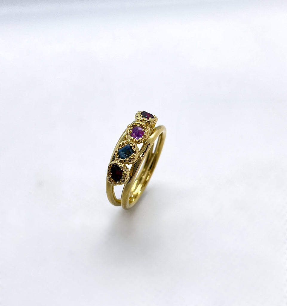 A ring stands up on its end. It is set with four uncut precious birthstones: a garnet, a sapphire, an amethyst and a ruby.