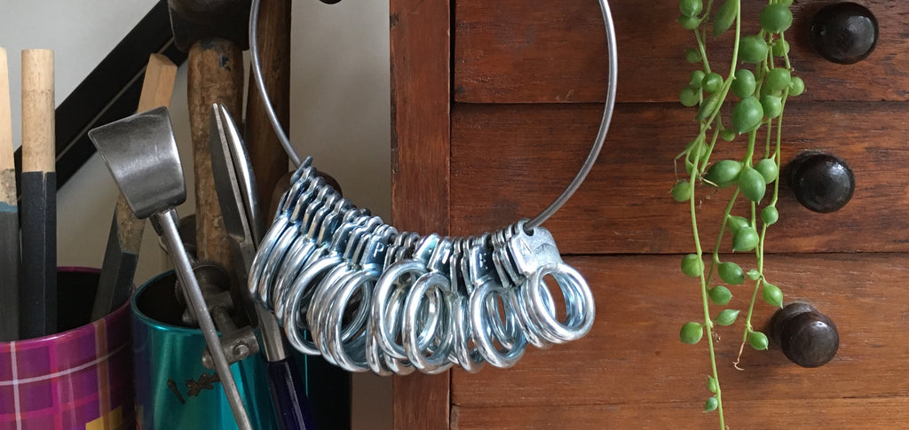 a metal loop of ring sizes hangs off a wooden cabinet. There are jewellers' tools to the left, and to the right a houseplant trails down.