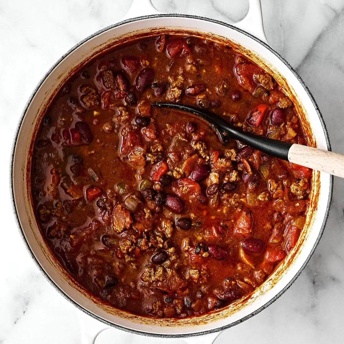 Pot of chili with beans and meat, with a ladle on a marble surface.