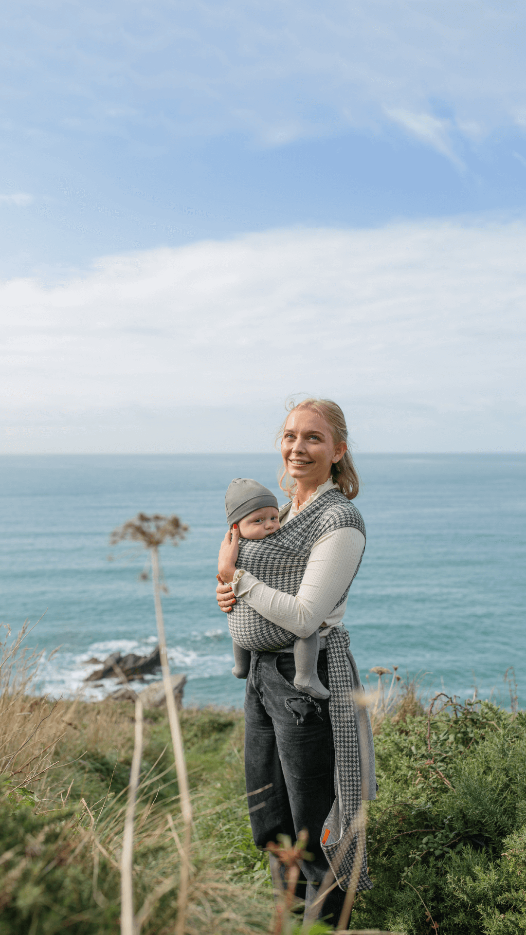 Mother smiling while holding baby in the Stormy Houndstooth Solly Wrap with blue skies and ocean in the background