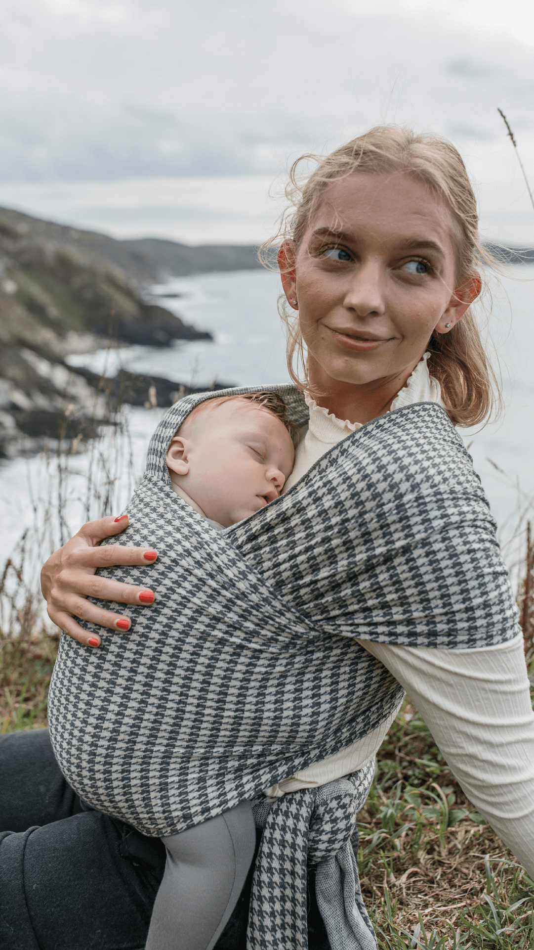 Mother glancing away while holding baby in the Solly Wrap in Stormy Houndstooth on the coast