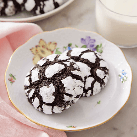 2 chocolate cookies with white powdered sugar sprinkled on top on a plate with a glass of milk