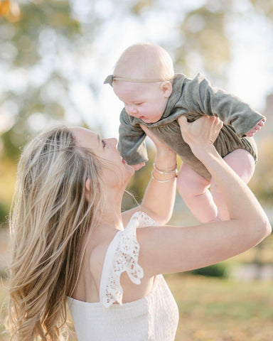 Blond woman holds baby in the air, outdoors and smiles