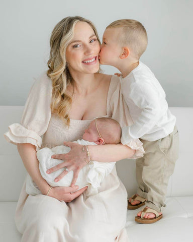 Blond woman sits on couch holding her baby, while young son stands and kisses her cheek.