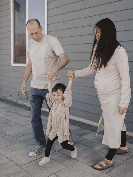 White man and pregnant, asian woman, each hold the hand of their small daughter and swing her