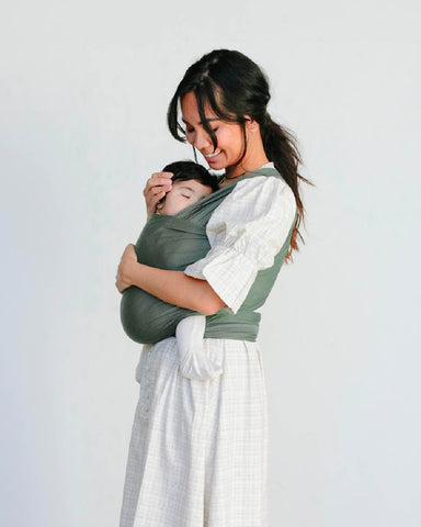 Woman with long black hair holds and looks down at sleeping baby in Basil Solly Wrap
