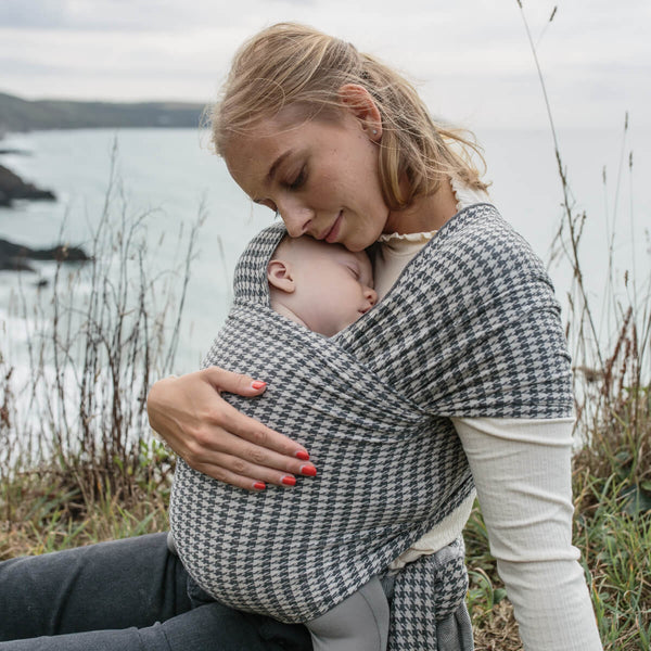 Mother lays head on sleeping baby in Stormy Houndstooth Solly wrap on English coastline