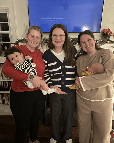 3 women stand together will arms around each other and smile. The women on the left and right are holding babies with their free arm.