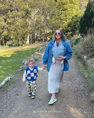 Woman holds hand of young son and walks on nature trail while wearing baby in Coastal Solly Wrap