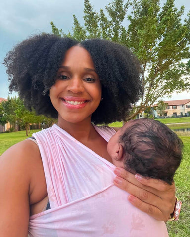 Woman with medium dark skin tone, and black curly hair smiles while babywearing in the Bloom Solly Wrap