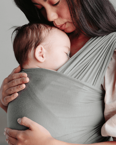 Mother leaning head down to kiss sleeping baby's head while he sleeps in a Mist Solly Wrap
