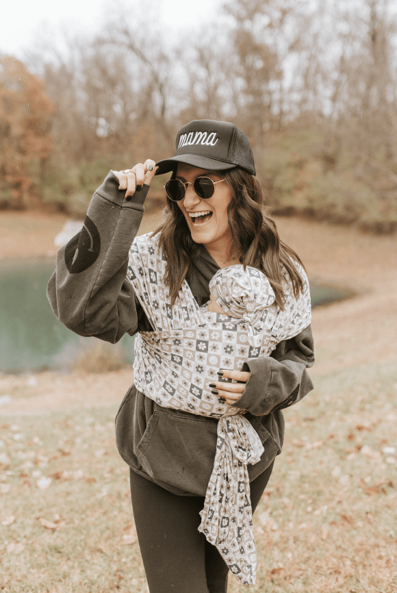 Woman with light skin town and long brown hair smiles while holding baby in Good Vibes Solly wrap at a park