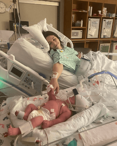 Mother in hospital bed smiles and reaches towards baby in a separate bed. Baby reaches back and they hold hands.