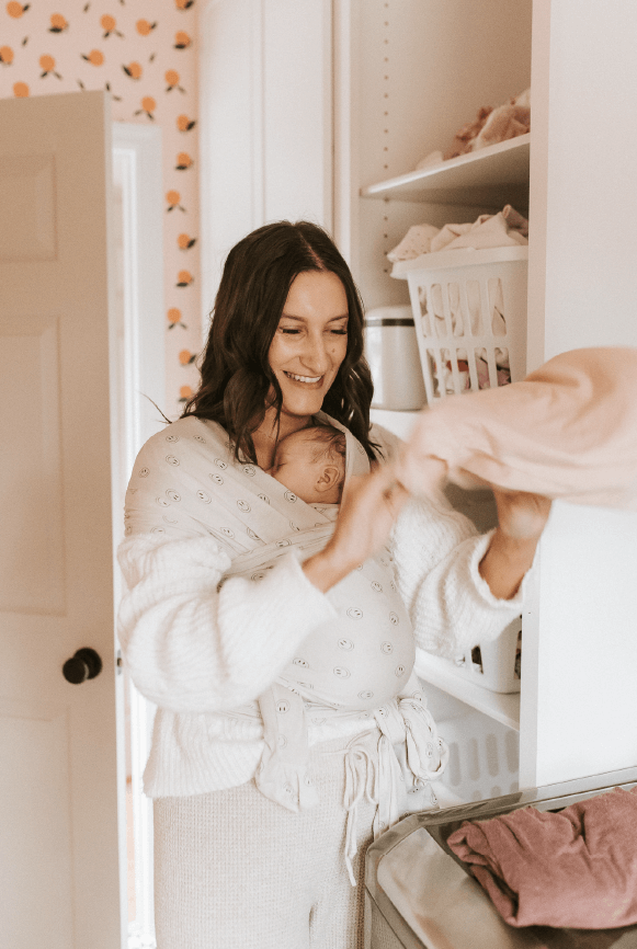 Woman with light skin tone and long brown hair smiles while folding laundry, wearing her baby in the Happy Days Solly Wrap