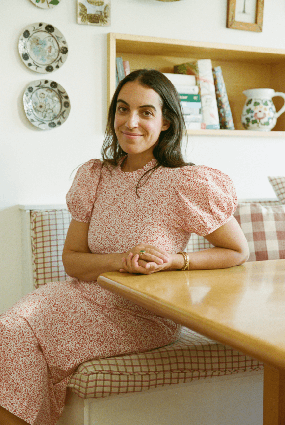 Brunette woman sits in a cozy kitchen nook and smiles in a floral dress.