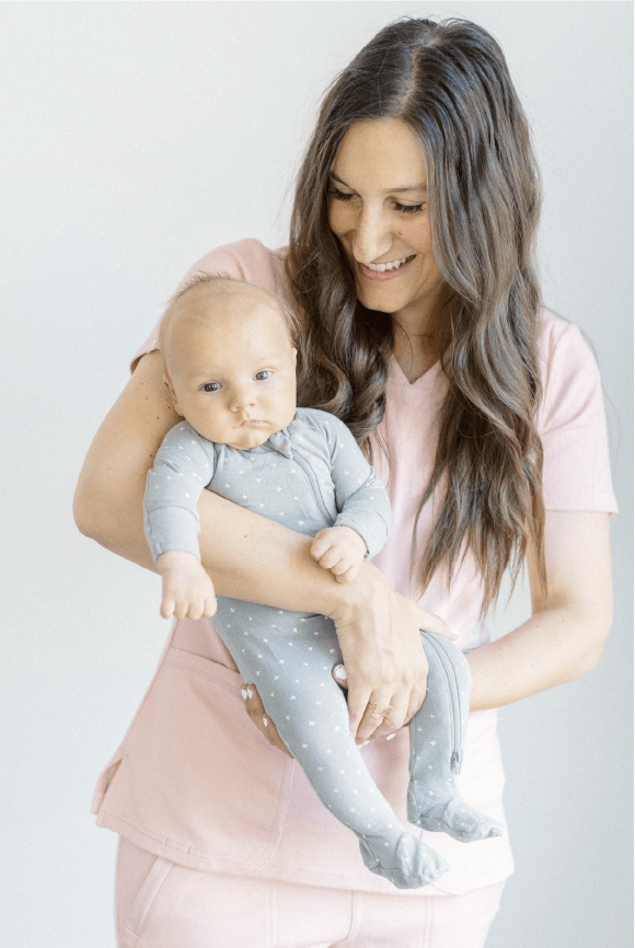 Woman with light skin tone in pink nurse scrubs, holding baby in gray sleeper