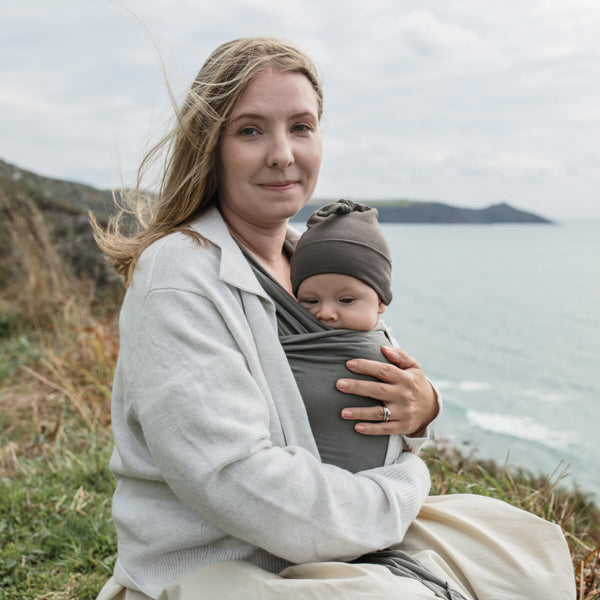 Blond woman holds baby in Sea Stone Solly Wrap, wrapped in a cardigan on the English Coast