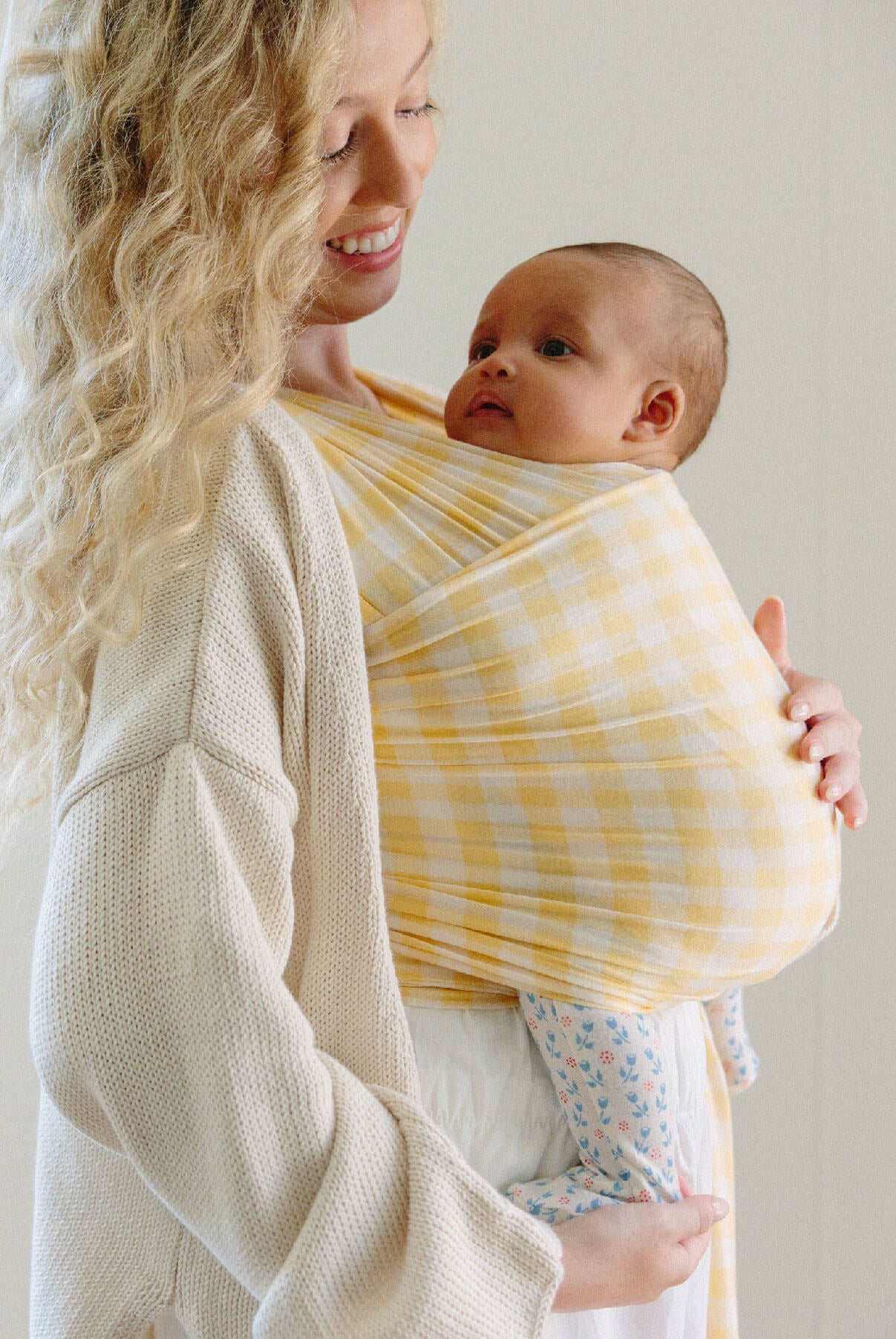 Mother with blond wavy hair smiles down at baby in a yellow gingham Solly wrap