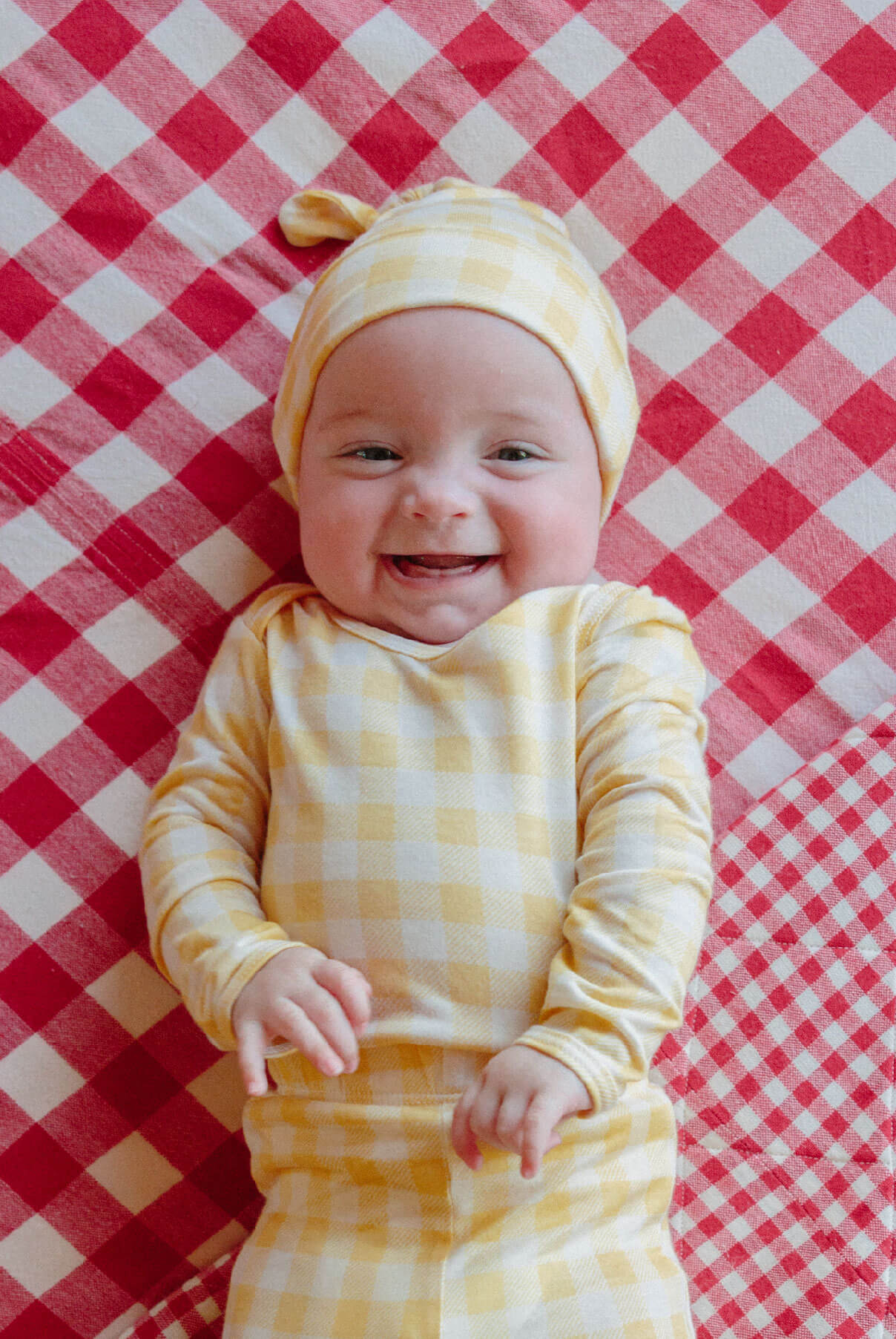 Smiling baby in a matching yellow gingham onesie and hat lies down on a red gingham blanket