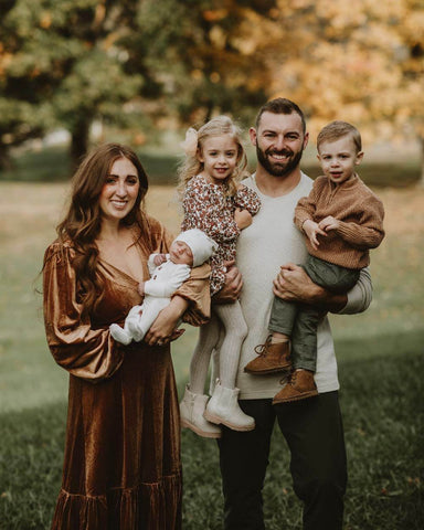 Family poses for picture in park with fall leaves. Mom holds baby, dad holds young daughter and toddler son