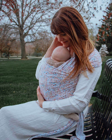Woman sits on park bench wearing baby in a floral Solly Wrap