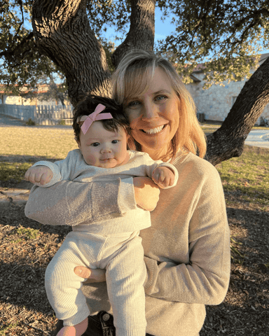 Blond woman holds up baby with black hair outside and smile