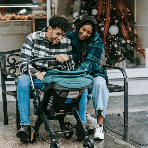 Couple with medium dark skin tone sit on street bench and smile at baby in stroller