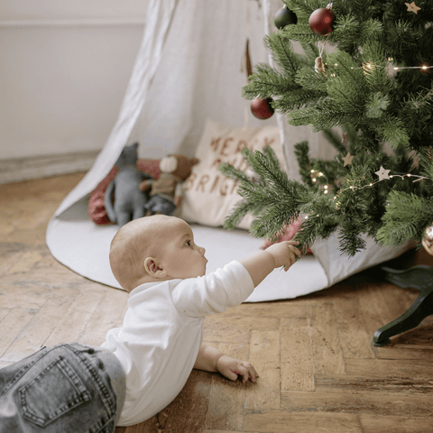 Baby with light skin tone lies on belly and reaches to the lowest branch of a decorated Christmas tree