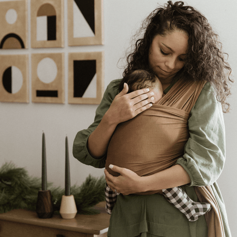 Mother with medium skin tone and curly brown hair looks lovingly down at baby in Camel Solly Wrap