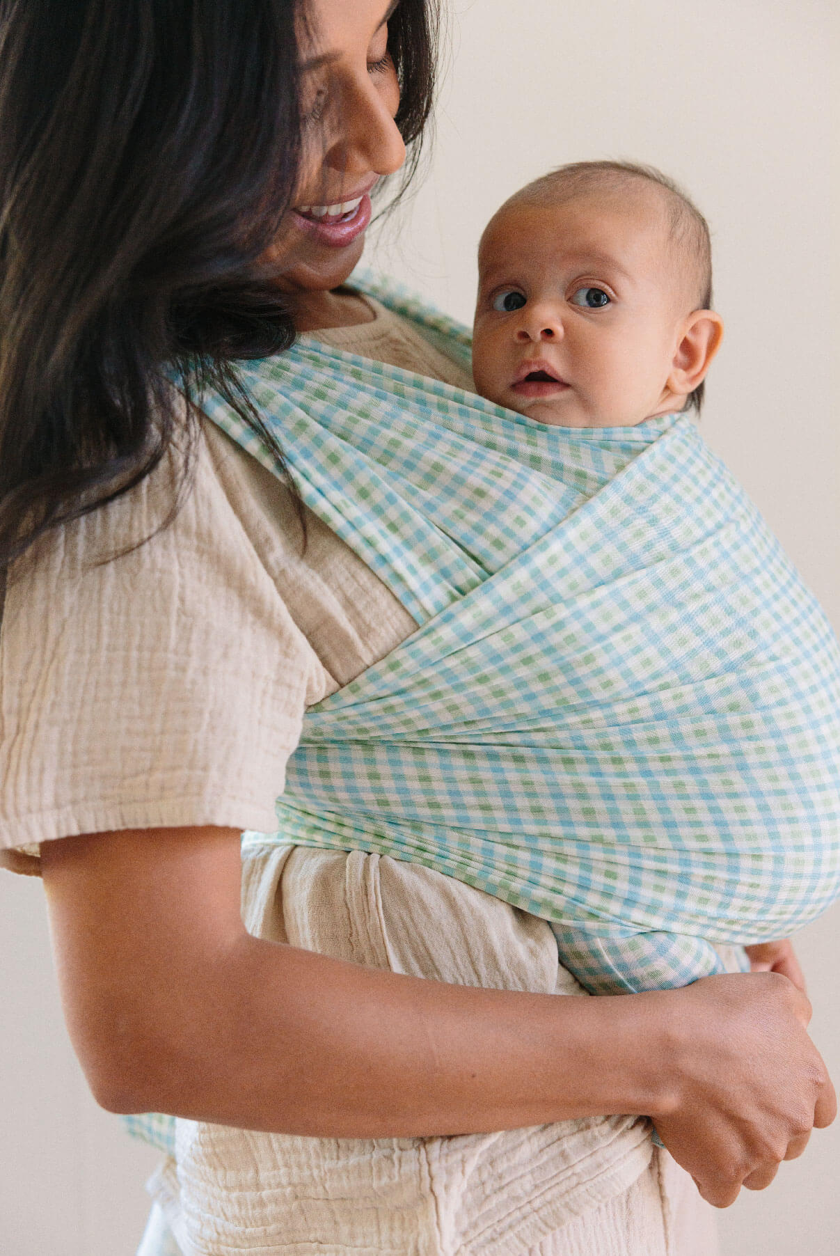 Brunette mother looks down at her baby wrapped in a green and blue gingham Solly wrap