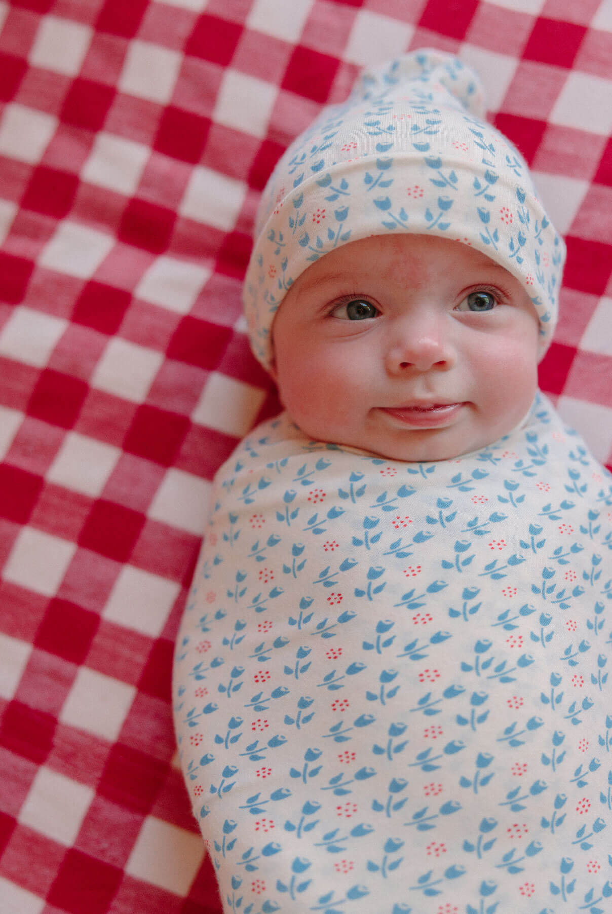 Baby smiles and looks off to side swaddled in "poppy" floral print on a gingham blanket