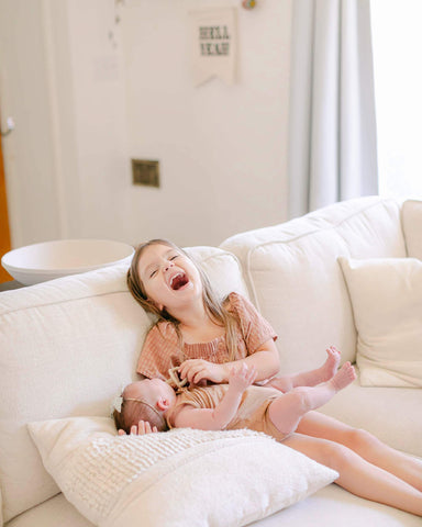 Young girl laughs with head thrown back as she sits on a couch with her baby sister in her lap