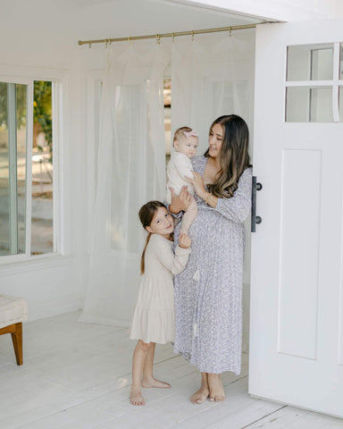 Woman stands holding and looking at baby while another child hugs her legs below