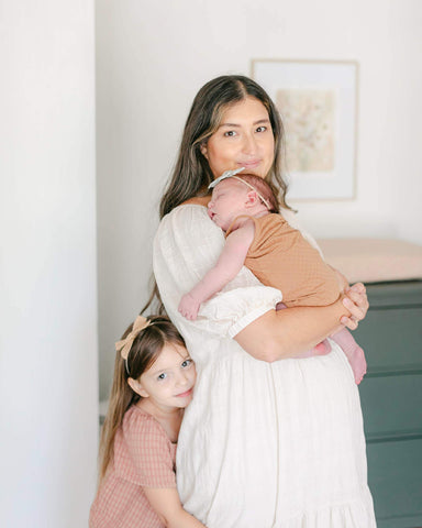 Woman with long brown hair smiles while holding baby and her young daughter hugging her legs