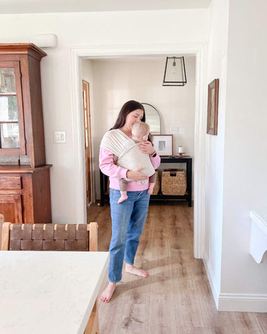 Woman stands in living room while holding baby in Flax Solly Wrap